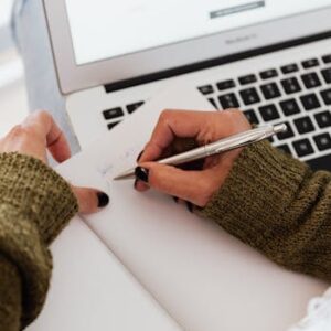 From above crop unrecognizable female in casual warm sweater taking notes in opened notebook with stylish silver pen while using modern netbook